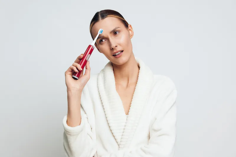 A young woman in a white bathrobe holds an electric toothbrush near her forehead with a confused expression. The plain background emphasizes personal hygiene and morning routine concepts.