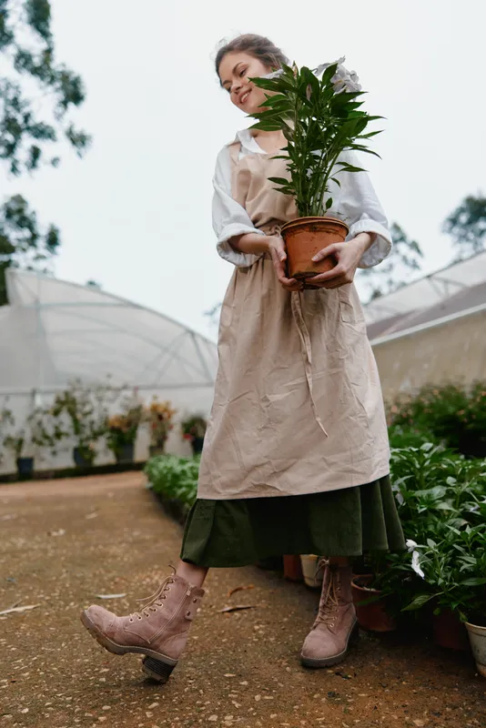 Young woman in an apron holding a potted plant, smiling in a greenhouse filled with greenery, conveying joy and a connection to nature, perfect for gardening concepts