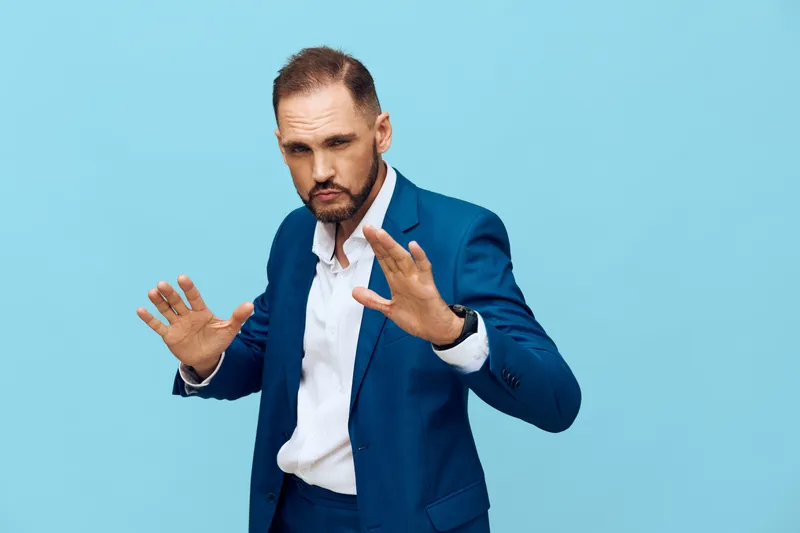 A professional man in a blue suit stands against a solid blue background, projecting a business mindset with calm authority, readiness, and measured focus in a boardroom ambiance.