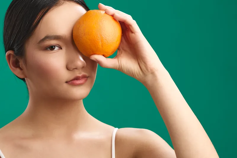 asian woman portrait in a studio setting holding an orange near her eye against a vivid green backdrop, creating a bold fashion and beauty mood that conveys modern confidence and elegance.
