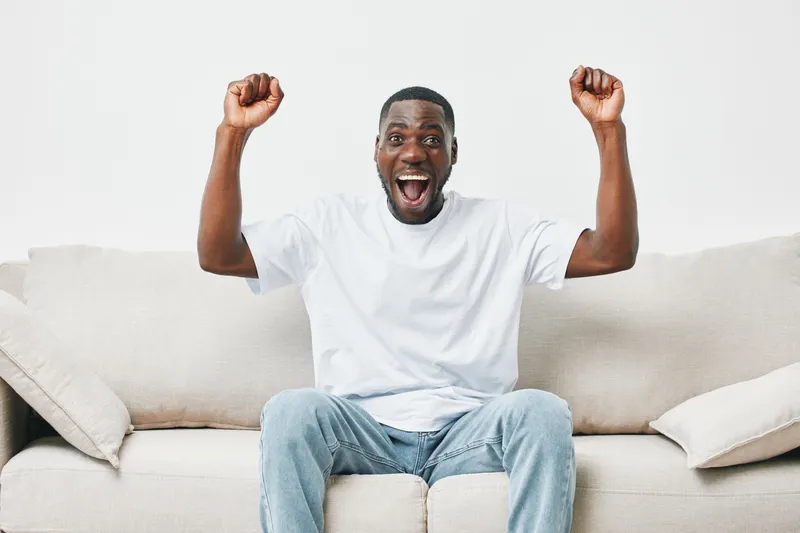 Excited young Black man celebrating success while sitting on a couch in a bright, minimalist living room, showcasing joy and positivity