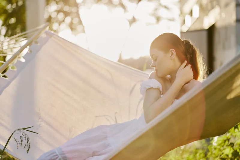 Serene young woman in a white dress relaxing in a hammock during sunset, evoking tranquility and connection with nature, highlighting summer vibes