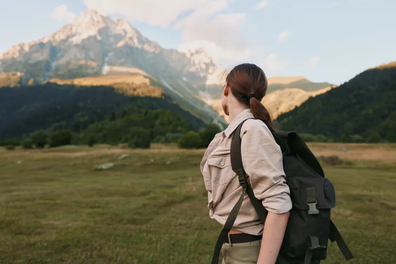 Candid woman outdoors, facing mountainous landscape, backpack ready, sunlit valley, casual attire, calm mood, natural colors, wide open scenery, sense of exploration