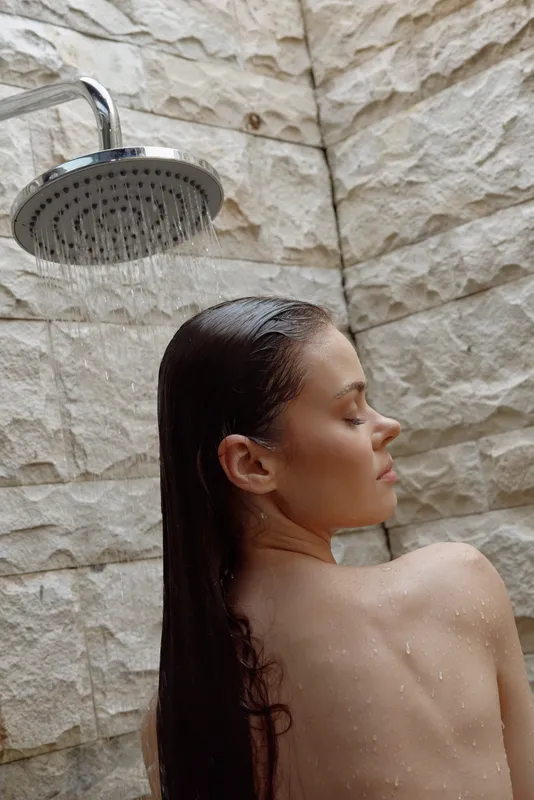 woman showering outdoors with wet hair against natural stone wall background, relaxed skin care moment in warm daylight, health and wellness concept