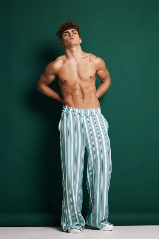 Young muscular man posing shirtless with striped pants against green background, showing fit body and confident expression in studio lighting.