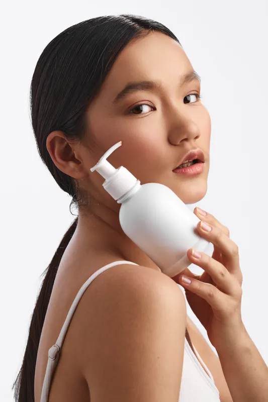 asian woman poses with a skincare bottle near her face in a clean studio, conveying calm confidence and self care through a minimal, modern aesthetic.