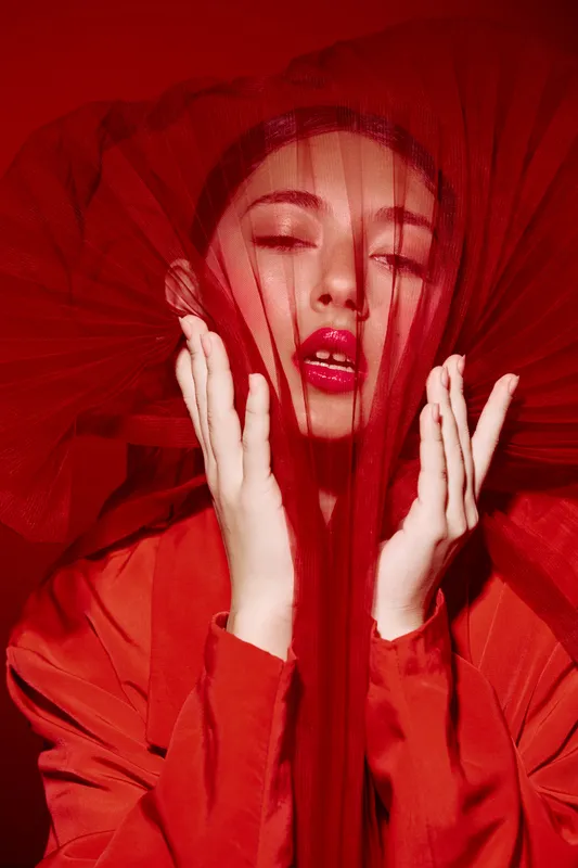 Fashion model in vibrant red outfit and tulle veil poses with hands near face. Close-up beauty portrait with dramatic makeup and styling. Concept for beauty and fashion creative.