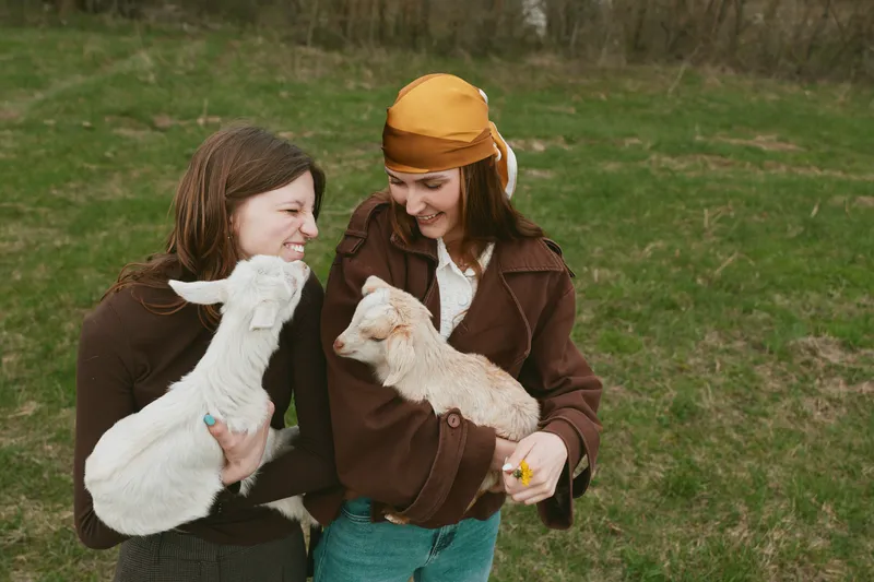 Two young women enjoy a sunny day outdoors holding baby goats while smiling and sharing a joyful moment in casual autumn clothing on a grassy field.