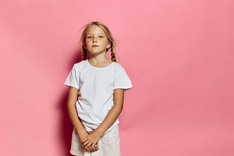 Young girl with blonde braids stands confidently in a white t-shirt against a pink background. Studio portrait with casual style and natural expression.
