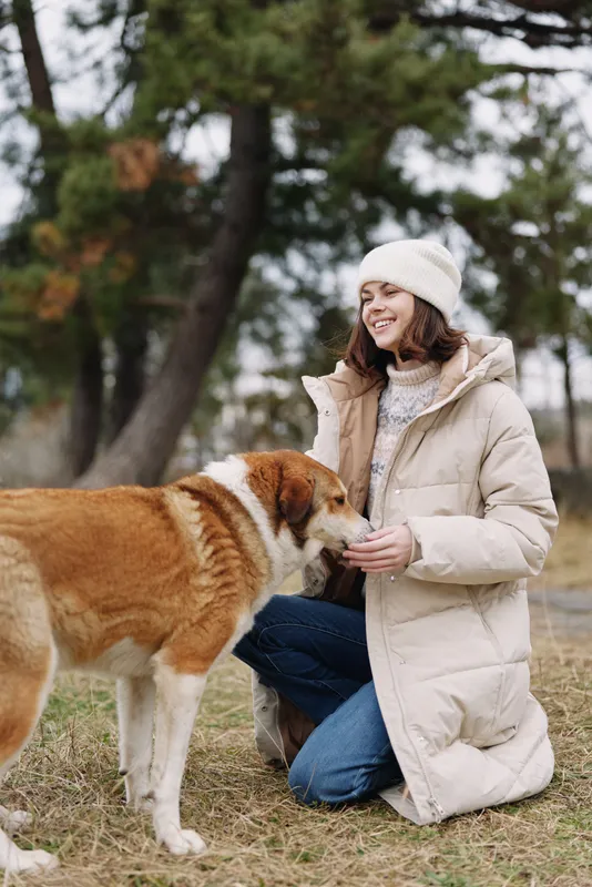Woman kneeling in a warm coat outdoors, smiling at a friendly dog during a chilly day in the park, showcasing tender interaction and genuine companionship.
