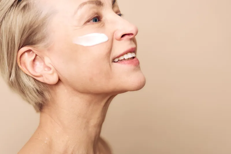 Smiling mature woman applies white face cream on cheek, showing skincare routine and healthy glowing skin. Concept of beauty, antiaging, wellness, selfcare, and natural skincare products.