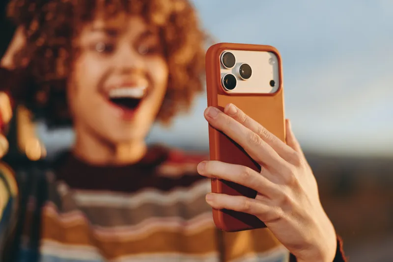 Woman makes a selfie with smartphone in hand, curly hair and smile in close portrait, lifestyle outdoors, blurred background, warm natural light, relaxed moment during golden hour outside