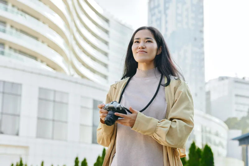 Camera in hand woman stands in an urban outdoor setting with modern buildings capturing moments during a travel day in the city