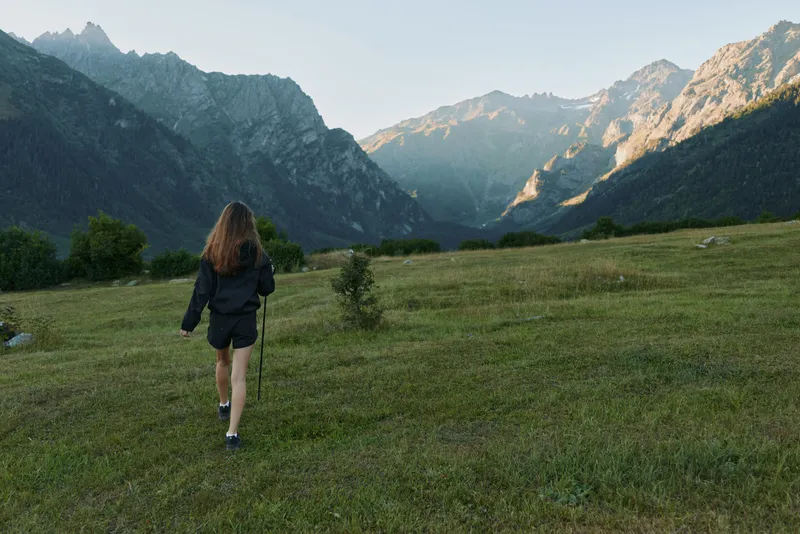 A lone hiker walks across an open meadow toward rugged mountains, creating a mood of adventure, exploration, and serene outdoor pursuit in a pristine alpine setting.