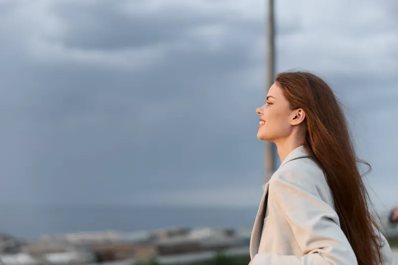 Confident woman in a business suit smiling by the seaside with a cloudy sky, representing professionalism and empowerment in a scenic setting.