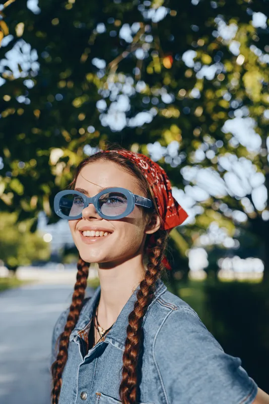 Lifestyle woman in modern boho-western denim street style with braided hair and sunglasses outdoors, film color, smiling and enjoying sunny day in natural green background.