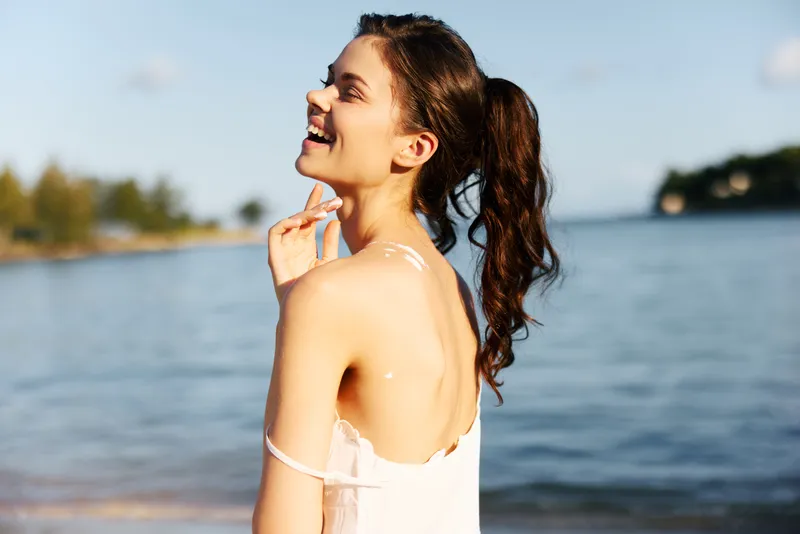 Portrait of a smiling young woman by the seaside, shoulder turned toward the camera, light breeze and sunlit skin, with calm water and distant shoreline in soft focus.