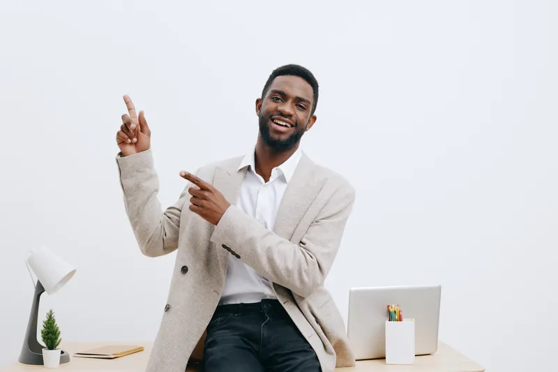 Confident smiling man with dark skin, beard, wearing beige blazer and white shirt, gestures with his hands while sitting at a modern office desk with laptop, plants, and stationery, studio background.