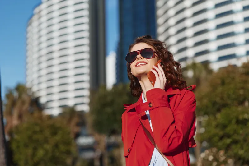 A stylish woman in a red coat talks on her phone in the bright urban outdoors, exuding confidence and chic city vibes under a clear sky.