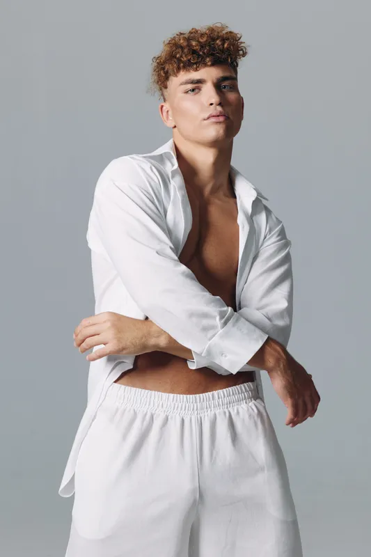 Young man with curly hair wearing white open shirt and white pants posing confidently against gray background in studio. Fashion, casual style and male model portrait.