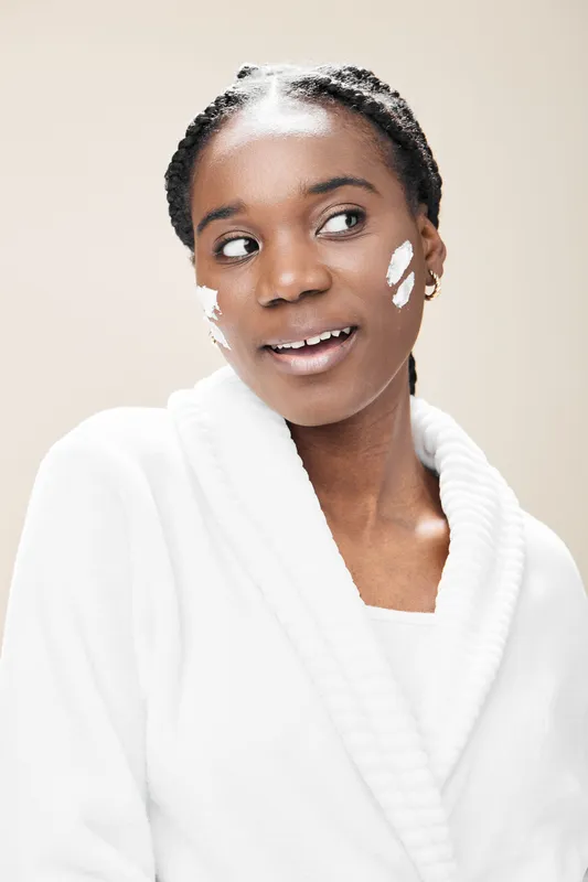 Young Black woman smiling with skincare cream on her face, wearing a cozy white robe against a soft cream background, conveying beauty and self care