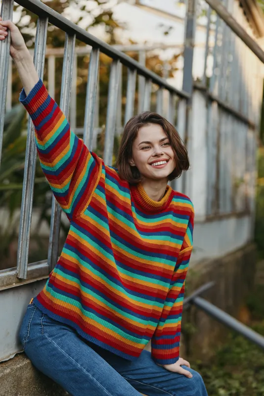 Woman smiling in a colorful striped sweater, casual portrait outdoors by a greenhouse frame, relaxed pose and natural light capturing a cheerful, stylish mood.