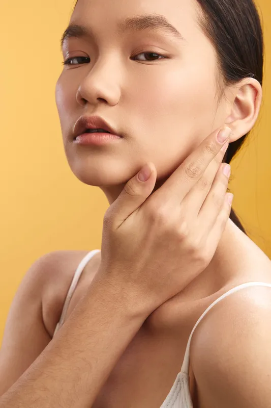 Asian woman with a calm expression gently touches her face in a studio portrait, highlighting skincare and beauty against a warm, soft backdrop that emphasizes elegance.