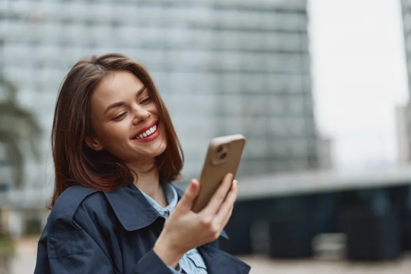 Young woman with brunette hair smiles at her smartphone outdoors in a modern urban setting, clearly enjoying a candid relaxed moment during a casual day