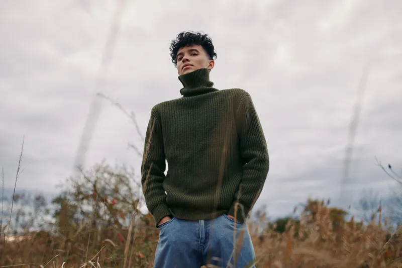 A solitary portrait of a young adult wearing a chunky green turtleneck and relaxed denim, conveying authenticity through natural posture and candid expression.