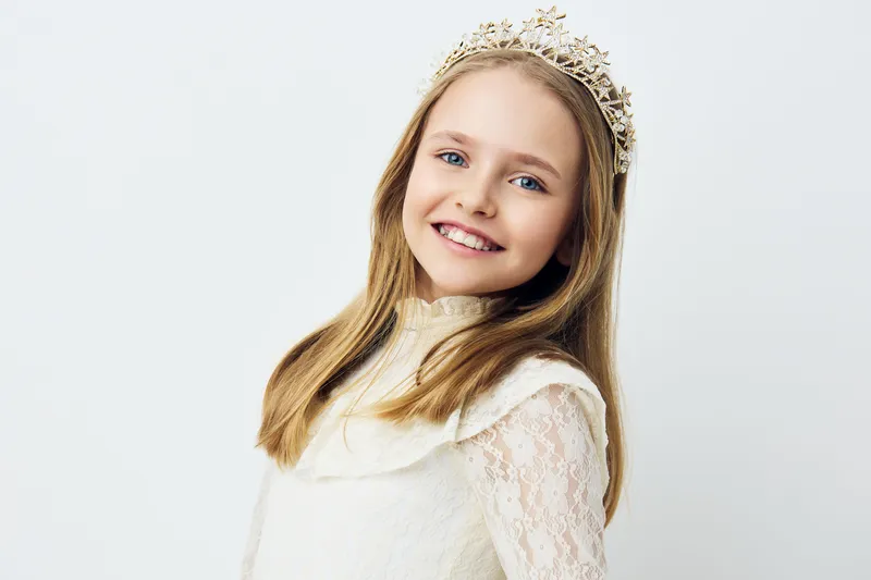 Smiling girl wearing a crown, dressed in a delicate lace dress, showcasing joy and innocence against a soft, light background