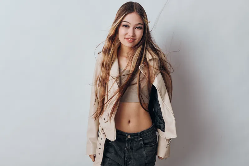 Young woman with long hair wearing casual jacket and crop top posing against white background looking at camera with confident expression and relaxed posture.
