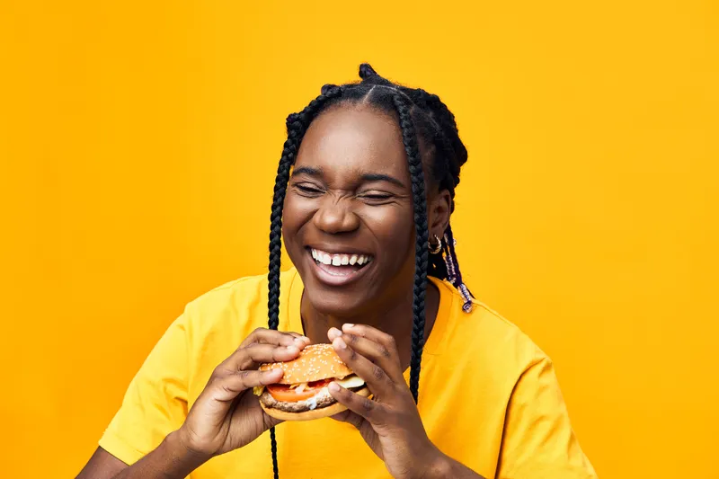 Joyful young woman enjoying a burger against a bright yellow background, wearing a cheerful yellow shirt, showcasing a healthy lifestyle and happiness in food
