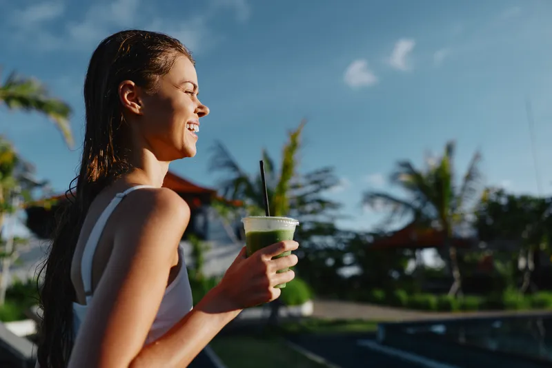 Cheerful woman outdoors, holding a green smoothie, smiling in sunshine by a pool, relaxed posture, tropical backdrop, vibrant colors, candid lifestyle moment, summer vibe