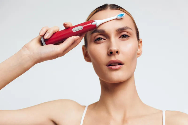 Young woman holding electric toothbrush near forehead with neutral expression on white background. Concept of dental care, hygiene, and personal grooming. Studio lighting highlights clean skin.