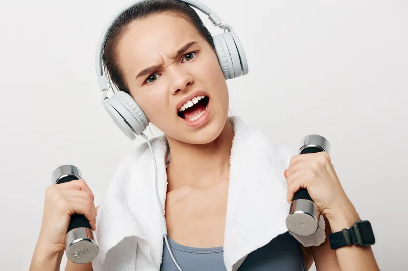 Athletic young woman in sporty tank top with towel around her neck, wearing wireless headphones and a smartwatch, lifting chrome dumbbells with a fierce determined expression on white background,