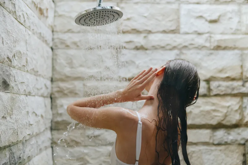 Woman taking outdoor shower with stone wall background, wet hair, and relaxed posture under rainfall, natural light enhancing the refreshing moment in summer setting.