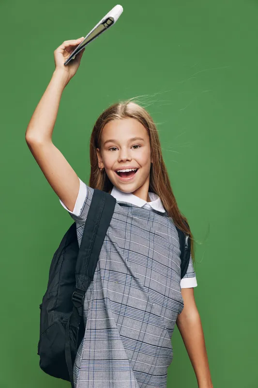 Girl student smiling and raising hand with book in backpack on green background. Happy schoolgirl showing readiness for class with excitement and education.