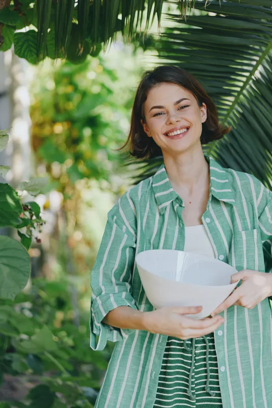 Smiling woman outdoors in a green striped jacket holding a white bowl, framed by lush foliage.