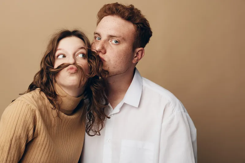 Young couple with playful expressions posing against beige background. Woman makes mustache with hair while man looks at camera. Concept of fun, love, and casual lifestyle.
