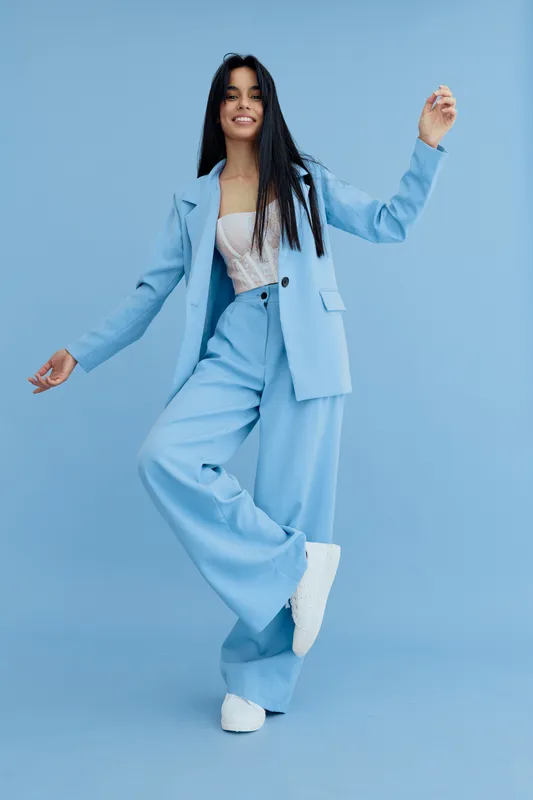 Young woman in blue suit dancing joyfully against a solid light blue background wearing white sneakers and a casual lace top, expressing happiness and movement.