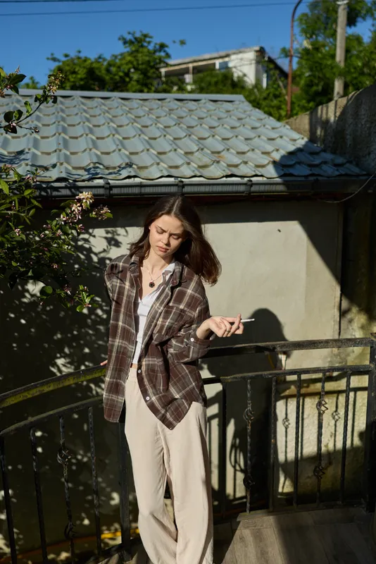 Candid woman outdoors, relaxed lifestyle on a sunlit balcony, wearing plaid shirt and light trousers, casual pose, natural colors, soft breeze, urban backdrop, sunlit breeze confident vibe