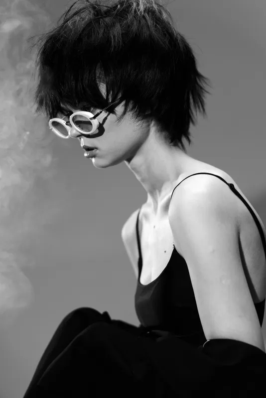 Black and white portrait of a woman with short hair wearing sunglasses and a black top, expressing a thoughtful mood and modern style.