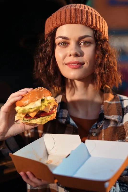 Woman with beanie and plaid jacket holding burger and takeout box, portrait outdoors in urban evening, casual smile and warm golden hour glow, authenticity, candid lifestyle and emotional