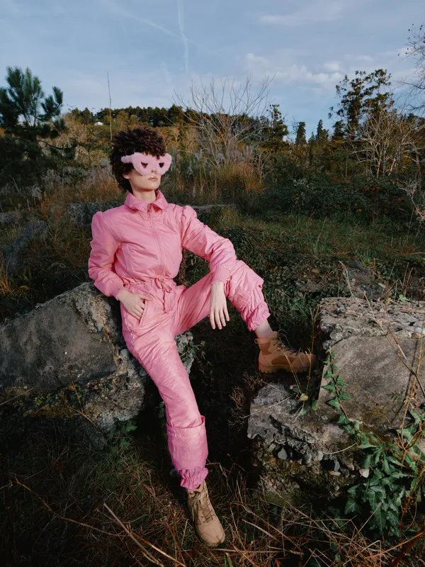 Pink jumpsuit fashion portrait outdoors perched on rocks in a natural landscape, wearing sunglasses and boots, showcasing bold style and relaxed confidence in a vivid rural setting.