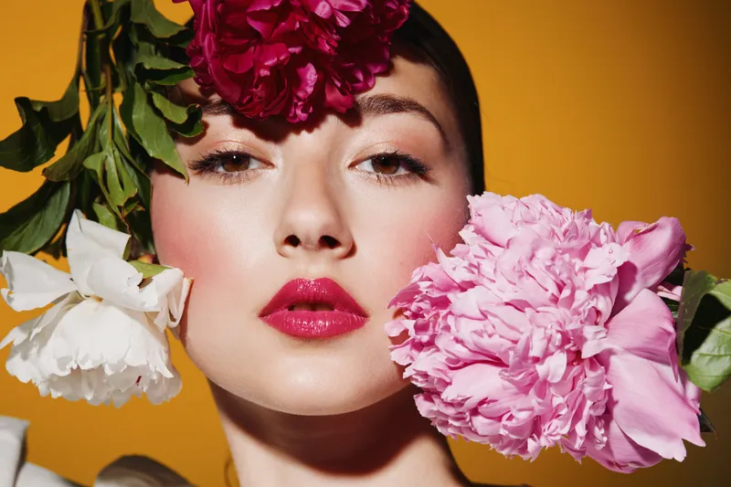 Portrait of a young woman with vibrant makeup and large pink and white flowers around her face. Creative beauty and fashion concept on a bright yellow background.