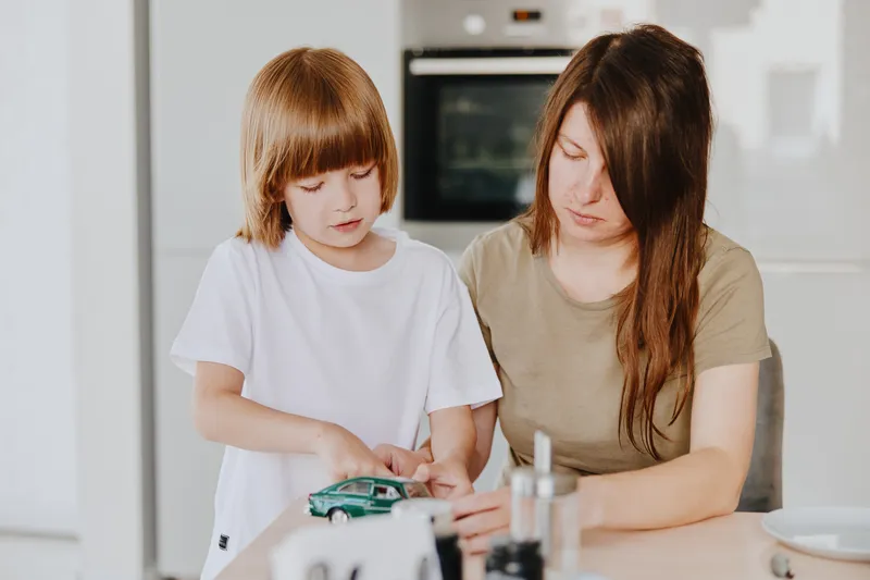 Caring woman playing with her son, engaging with a toy car in a bright, modern kitchen. A moment of interaction and bonding, showcasing family love and joy.