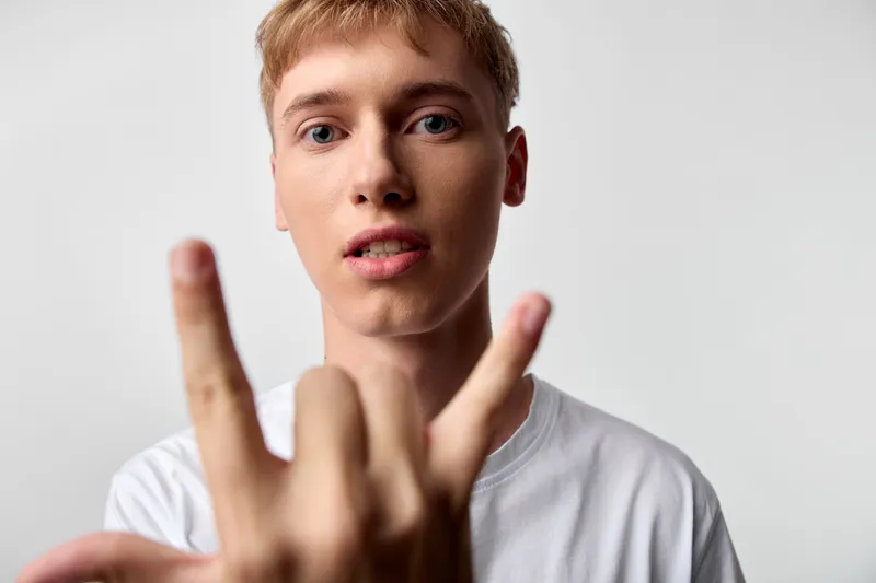 Close up of a young man making a rock on gesture with fingers, wearing a white tee, soft studio lighting, plain background, casual confident mood, modern youth expression