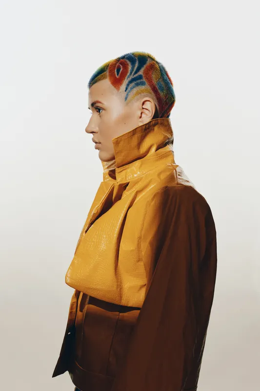 Woman with colorful shaved hairstyle wearing a shiny yellow jacket in profile, displaying edgy fashion and bold artistic expression with vibrant hair patterns.