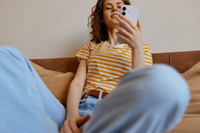 Casual woman lounging on a sofa at home, scrolling on a smartphone. Striped tee, denim, relaxed pose, warm light, lifestyle moment focused on comfort and self expression