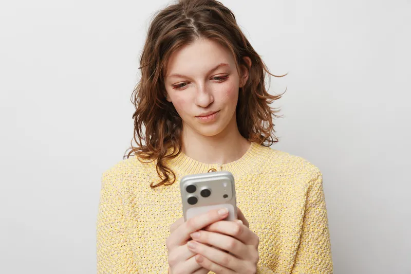 Young woman in yellow sweater looks at smartphone with slight smile, isolated on white background. Concept of communication, technology, social media, and casual lifestyle.